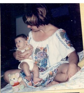 A young woman with short ash-blonde hair holds an infant with dark hair. The young woman's face is pointed away from the camera; she sits cross-legged and is dressed in a white dress covered in colorful embroidery.