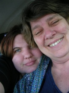 Two women smile at the camera. One is older, with short ash-blonde hair and a toothy smile. The other is younger, with a closed mouth smile and red hair. She rests her chin on her mother's shoulder, and it's clear she's holding the camera for both of them.